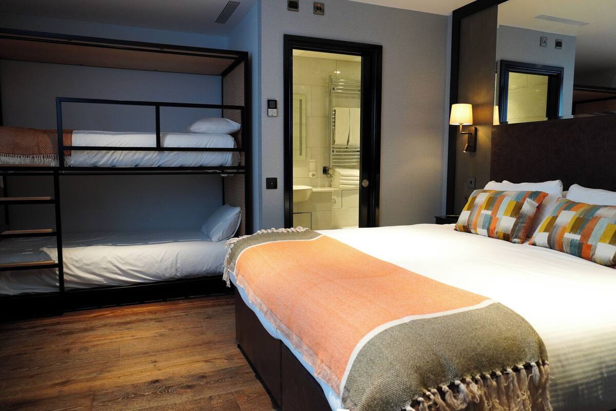 A hotel room with a bunk bed, a single bed, a bathroom visible through an open door, and a nightstand with a lamp.