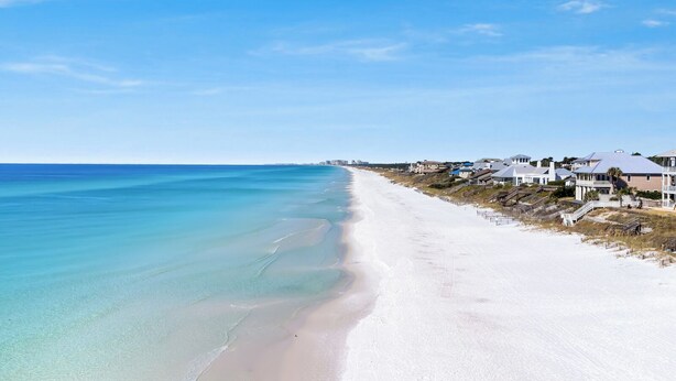 Une plage côtière avec du sable blanc, une eau turquoise et limpide, et des maisons dotées de balcons donnant sur l’océan.
