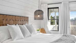 Une chambre à coucher avec une tête de lit en bois, du linge de lit blanc, un luminaire suspendu et une vue sur l’extérieur grâce à une grande fenêtre.
