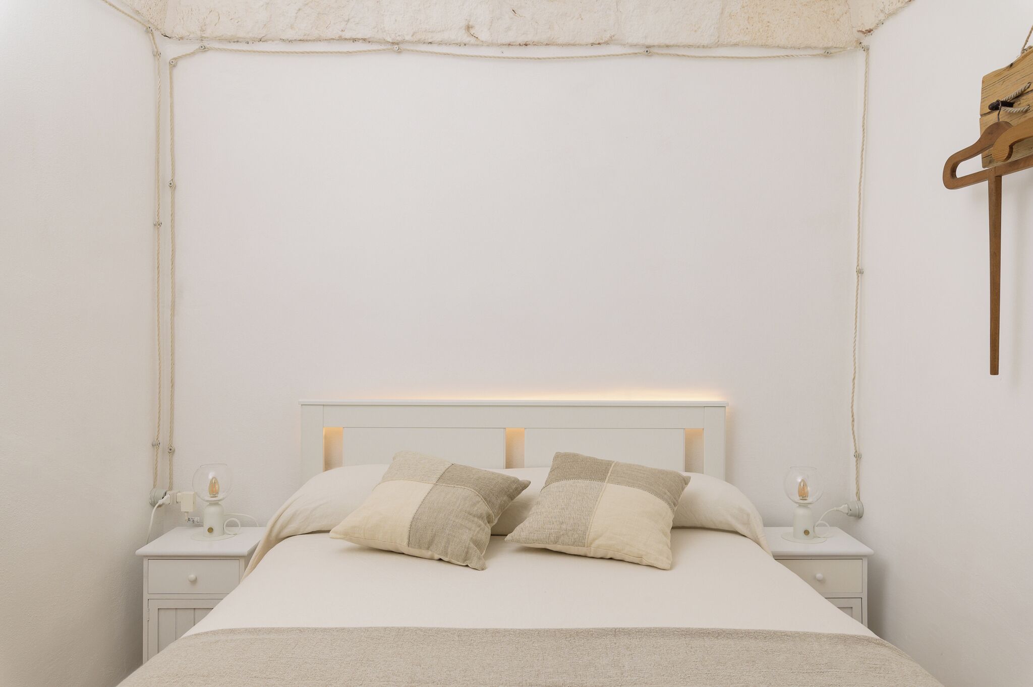 A neatly made bed with two pillows, a bedside table with a lamp, and a wooden hanger on the wall.