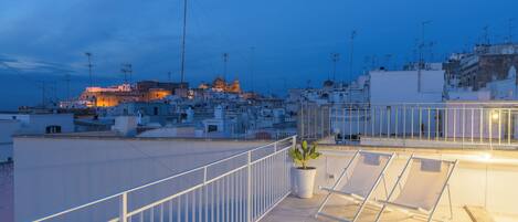 Terraza en la azotea con una planta en maceta y sillones de descanso, con vistas a un paisaje urbano que incluye edificios y una estructura destacada al fondo.