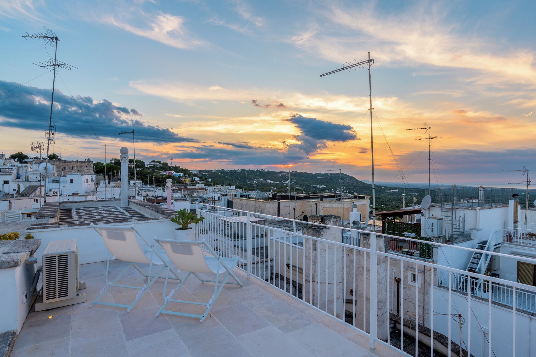 Une terrasse sur le toit, dotée de rampes blanches, d’un espace salon et offrant une vue sur le paysage urbain au coucher du soleil.