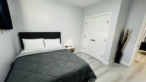 A neatly made bed with a dark quilt, a bedside table with a lamp, and a white wardrobe with a door handle. - Modern & Cozy 2 Bedroom Apartment in Seton, Calgary: Haven Away from Home! (Calgary)