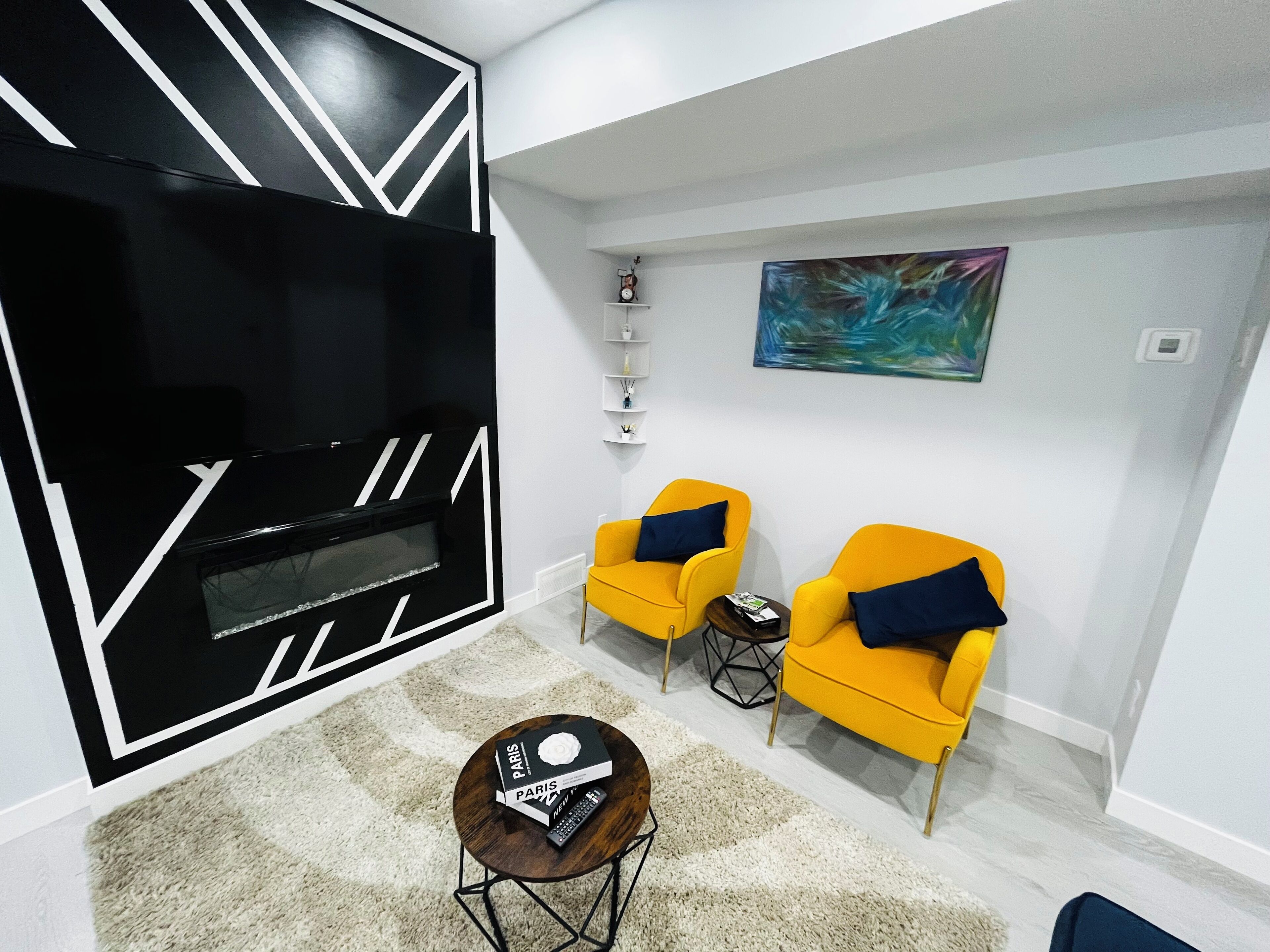 A modern living room with a black and white geometric wall design, two yellow armchairs with blue cushions, a small round coffee table, and a painting on the wall.