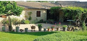 A stone building with a tiled roof, surrounded by a garden with potted plants and a grassy lawn.