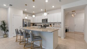 Una cocina moderna con armarios blancos, una isla central con taburetes de bar, electrodomésticos de acero inoxidable e iluminación colgante.