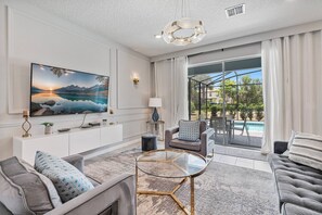 Uma sala de estar moderna com uma grande TV de tela plana, assentos confortáveis, uma mesa de centro de vidro e vista para uma piscina através de grandes portas de correr.