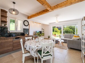 A modern kitchen with a dining area, a living room with a sofa, and a large window offering a view of greenery.