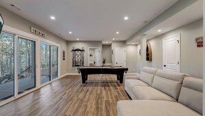 Amplia sala de estar con mesa de billar, un sofá color beige y ventanas grandes que ofrecen vistas al exterior.