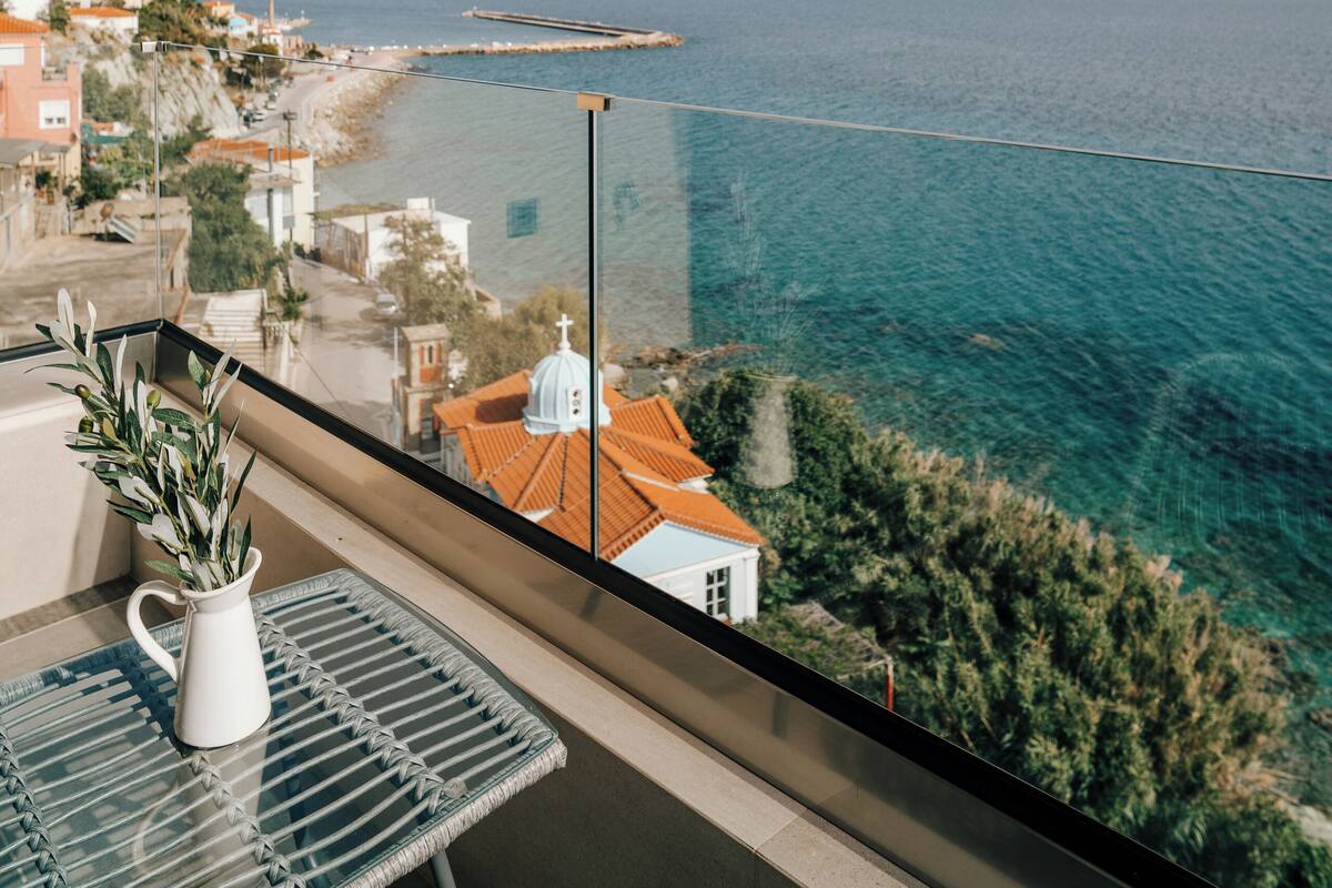 Un balcón con vista al mar, una jarrón blanco con plantas y un edificio con techo rojo.