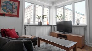 A modern living room with a sofa, a coffee table, and a TV on a wooden stand. There are three windows with potted plants on the windowsill. - Perfect located Apartment in the heart of Södermalm (Stockholm)