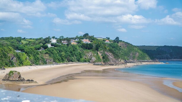 A coastal beach with a sandy shore, a rocky outcrop, and buildings on a hillside.
