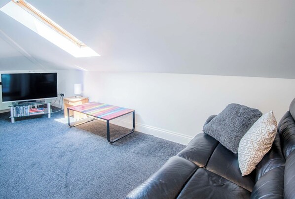 A modern living room with a black leather sofa, a colorful striped coffee table, a flat-screen TV on a stand, and a skylight above. - Rooftops - 1 Bedroom Apartment - Tenby (Tenby)