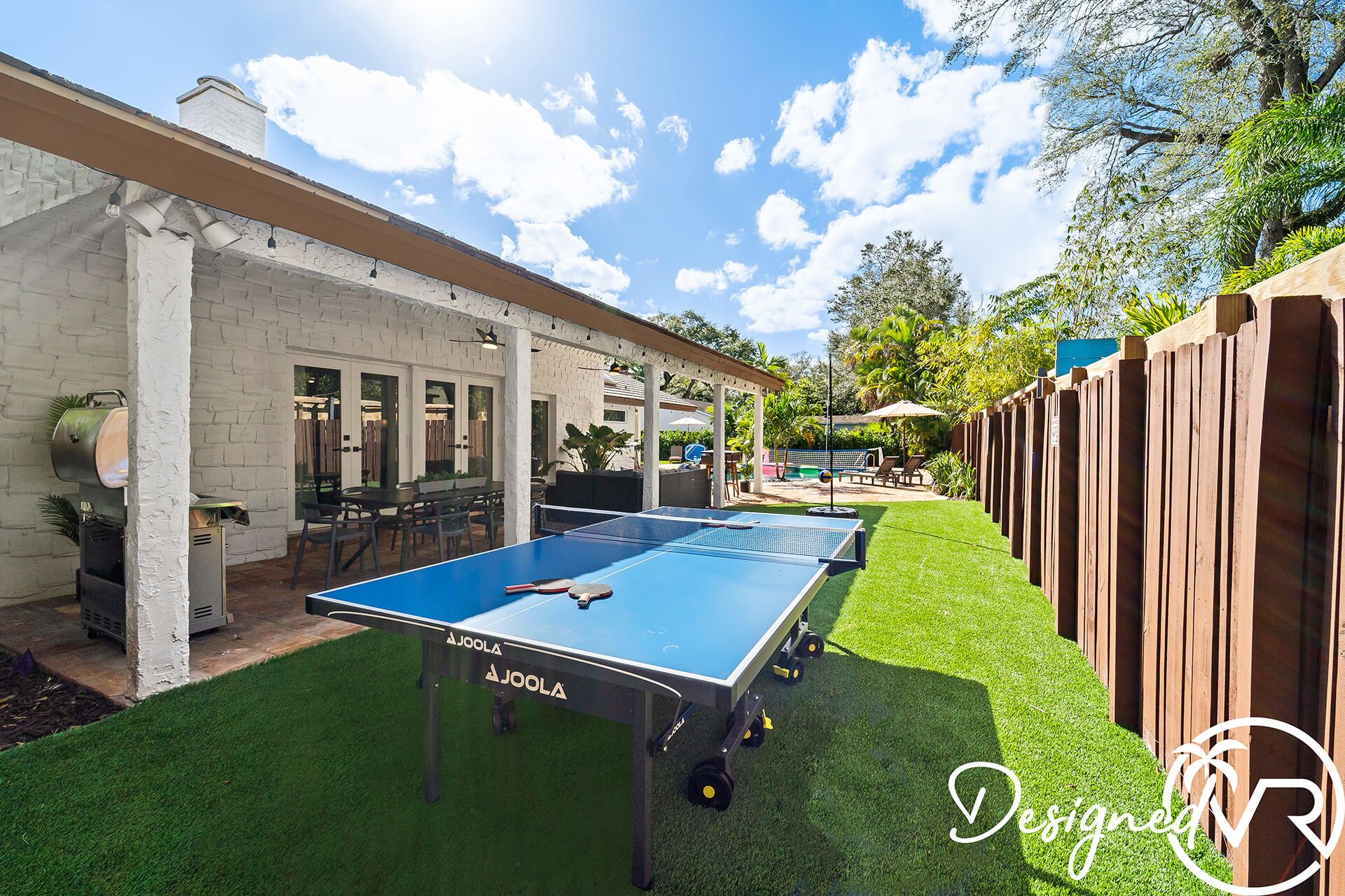 An outdoor ping pong table in a backyard with a brick wall, patio furniture, and a wooden fence.