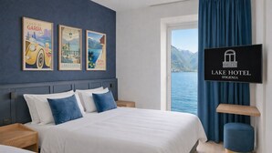 A hotel room with a bed, a nightstand, a TV, and a window with a view of a lake. - Lake Hotel Ifigenia (Nago-Torbole)