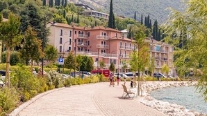 A coastal promenade with a row of multi-story buildings, a paved walkway, and a rocky shoreline. - Lake Hotel Ifigenia (Nago-Torbole)