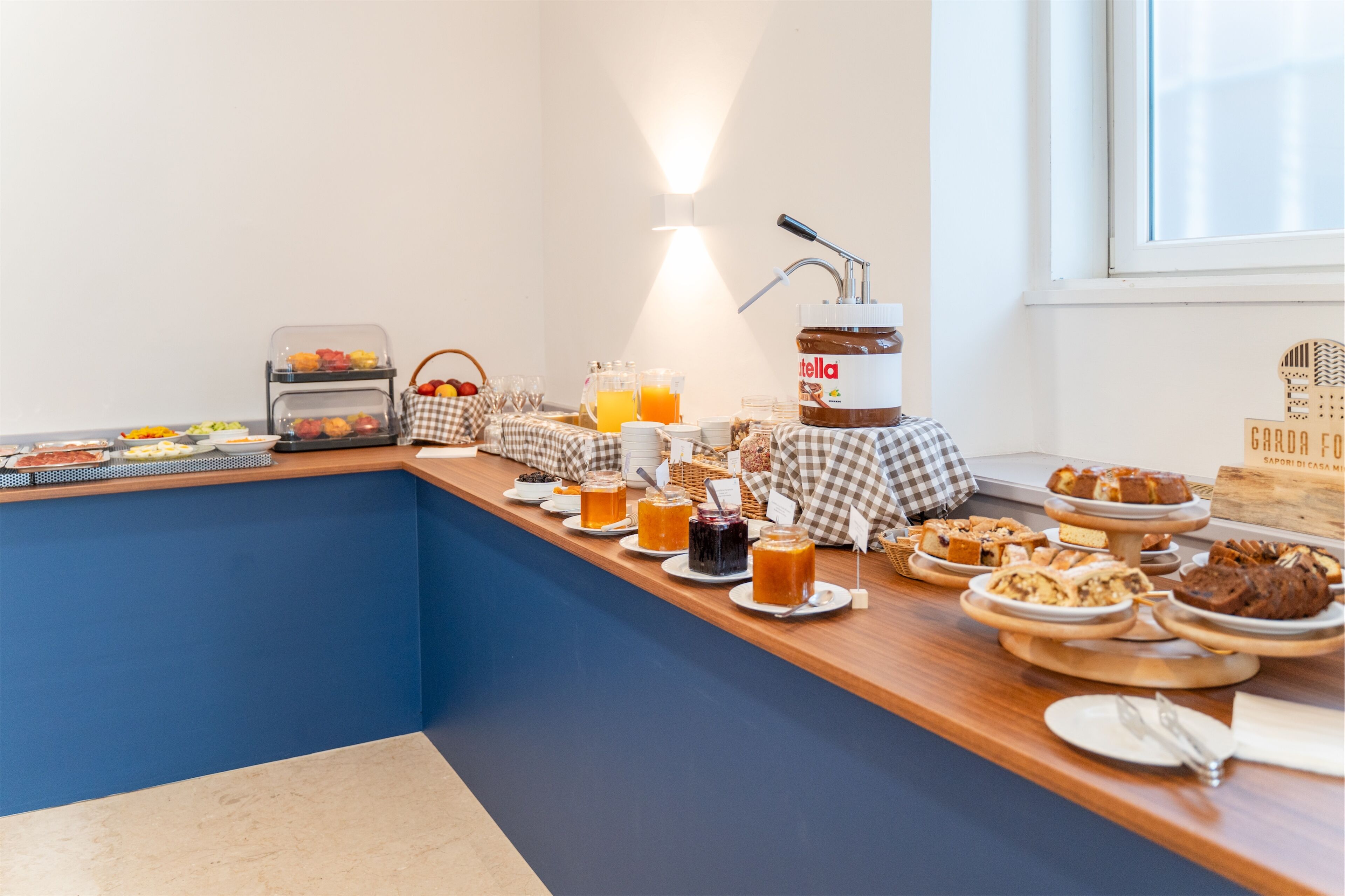 A breakfast buffet with various food items and drinks, a jar of Nutella, and a window with a view.
