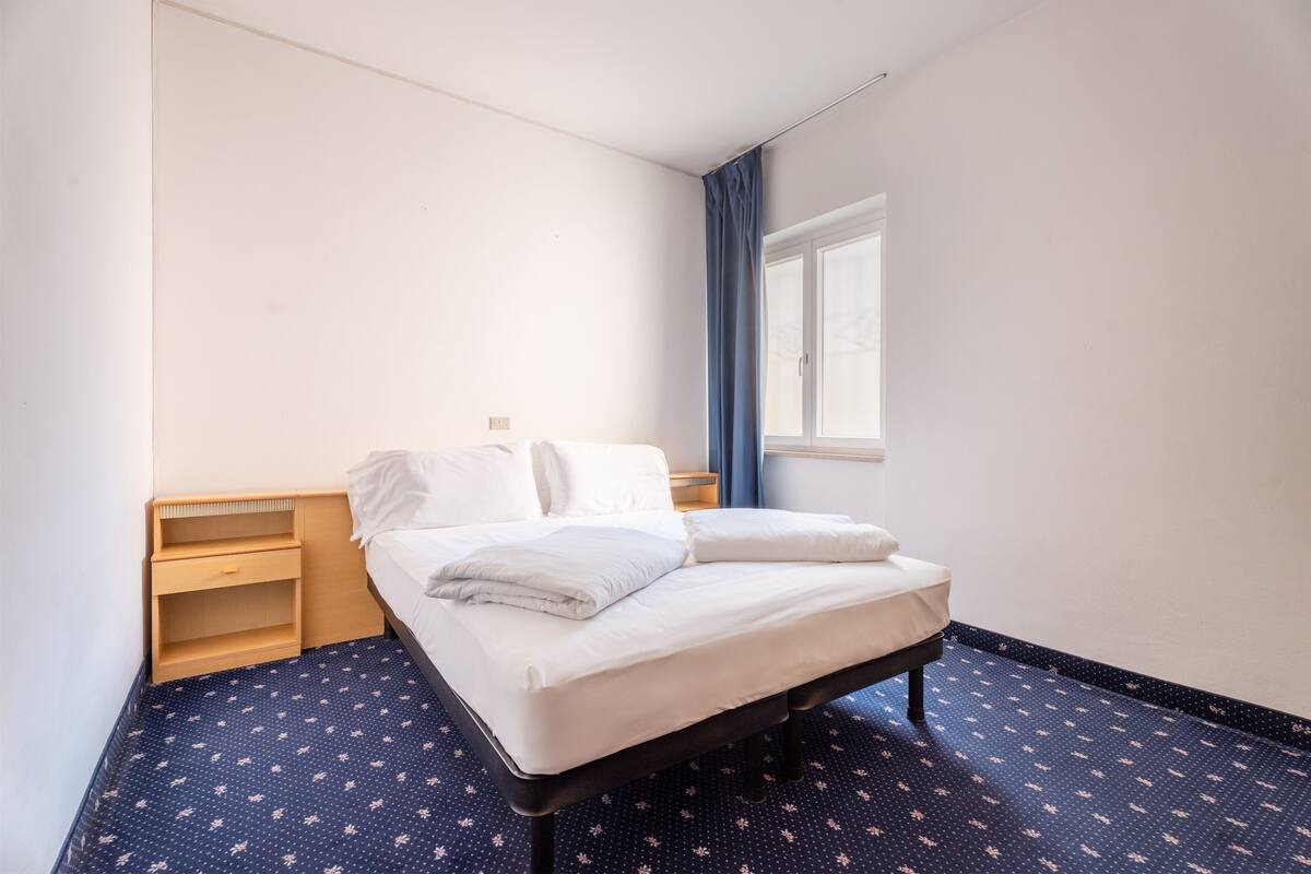 Un lit simple avec un matelas et des oreillers blancs, une table de chevet en bois avec un tiroir, une petite étagère et une fenêtre avec des rideaux bleus.