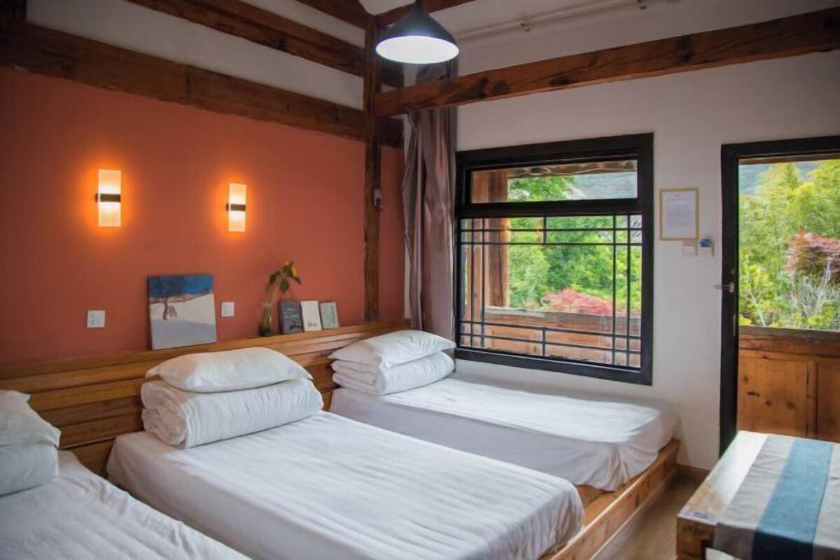 A room with two beds, wooden beams, a window with a view of greenery, and wall-mounted lights.