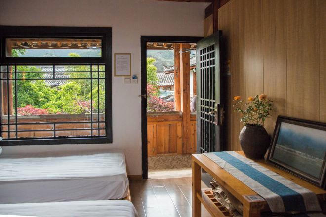 A traditional Japanese room with tatami flooring, a low wooden table with a vase and picture, and a sliding door leading to a balcony with a view of a traditional Japanese garden.