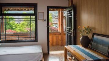 A traditional Japanese room with tatami flooring, a low wooden table with a vase and picture, and a sliding door leading to a balcony with a view of a traditional Japanese garden.