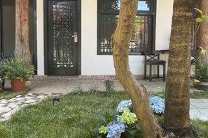 Una casa con puerta negra, un jardín pequeño con césped y flores, y un árbol en primer plano.