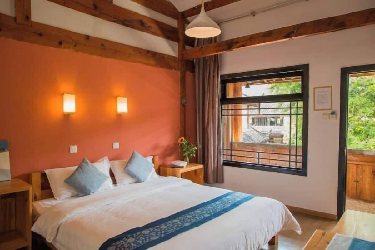A bedroom with a large bed, wooden beams, a window with a view, and a nightstand with a lamp.