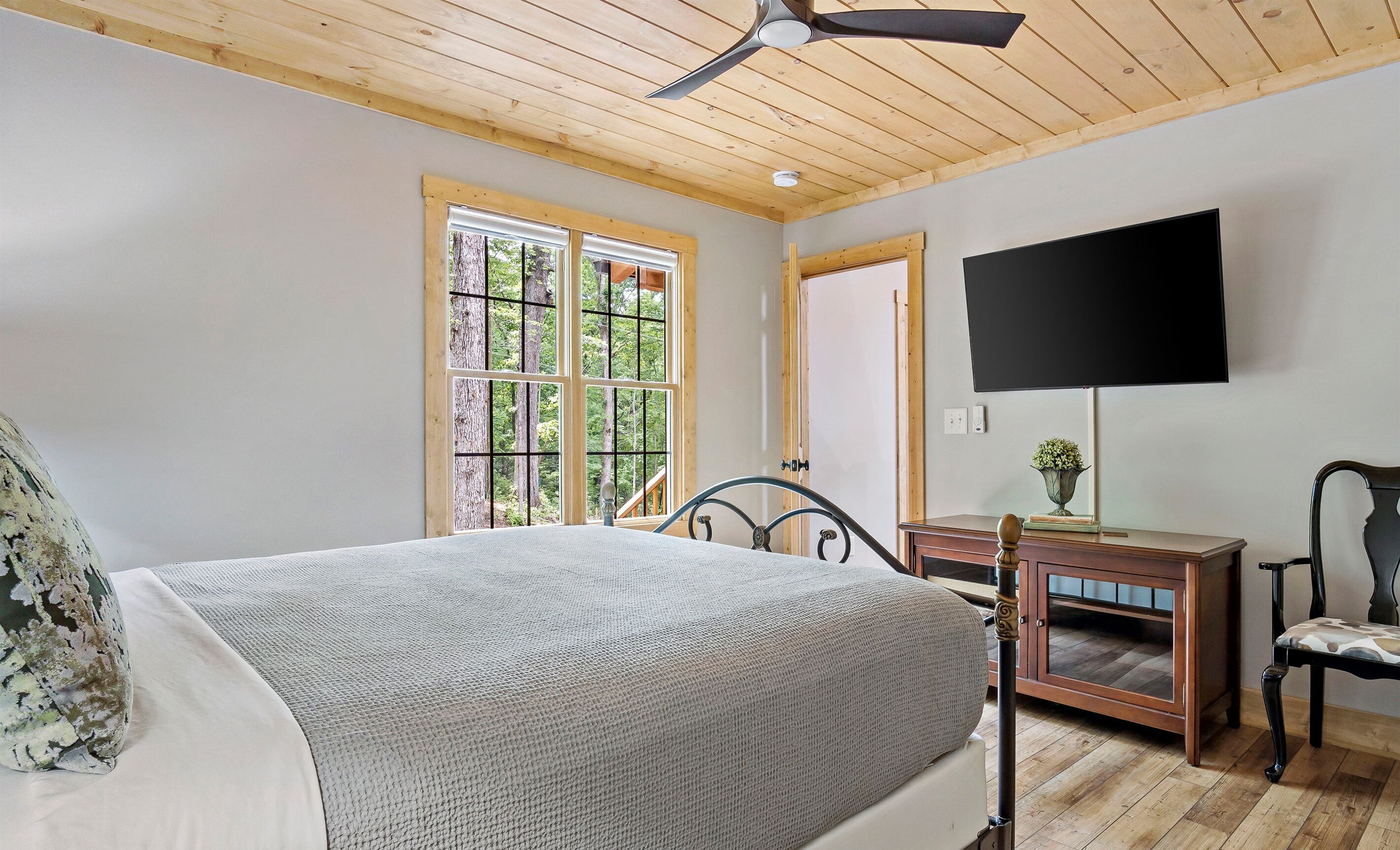 Un dormitorio con cama, escritorio, silla, televisión y ventanas amplias con vistas a árboles.