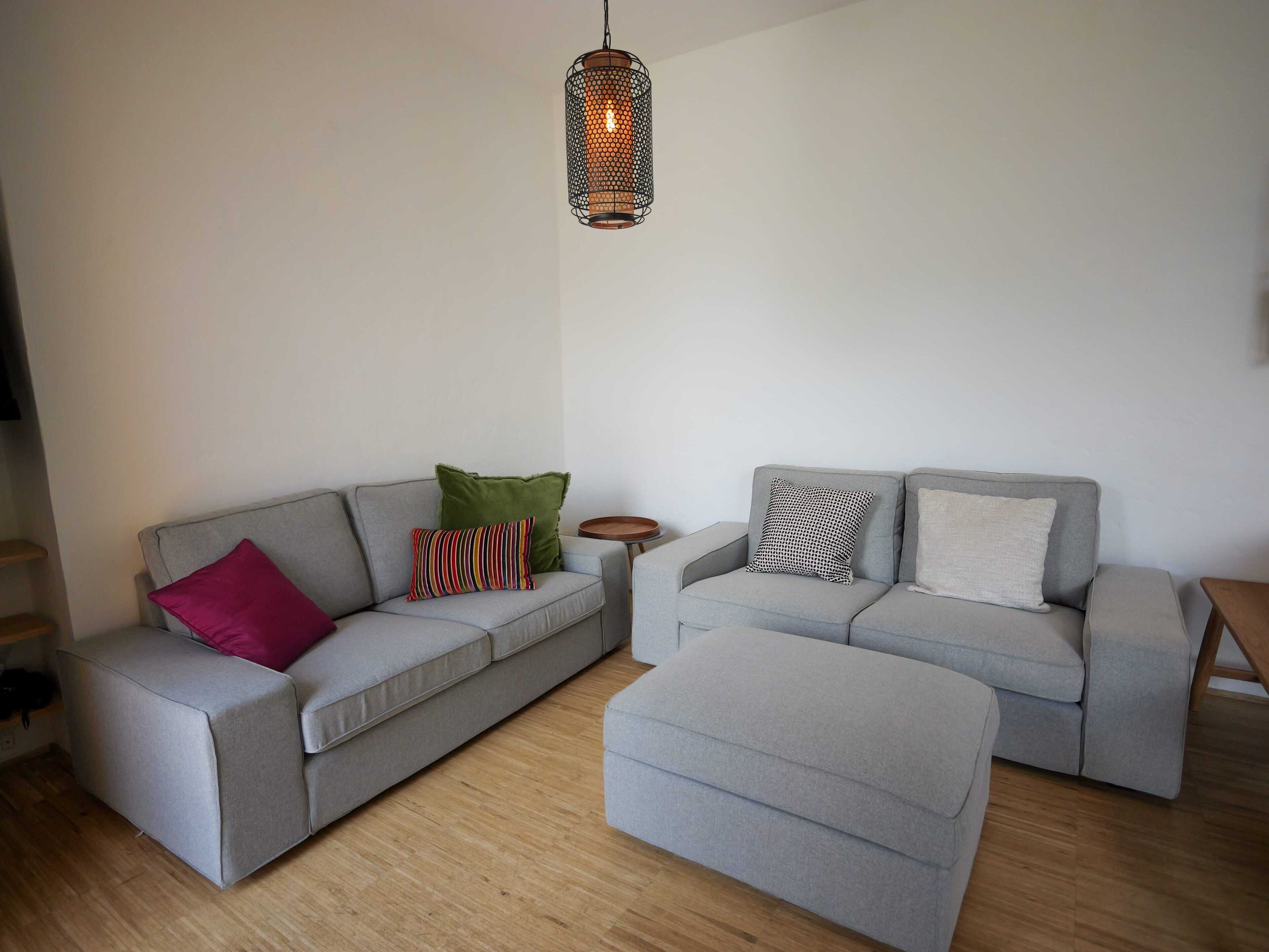 Una sala de estar moderna con dos sofás grises, uno con un cojín rojo y multicolor, y otro con cojines estampados. Se ve un piso de madera y una lámpara colgante.