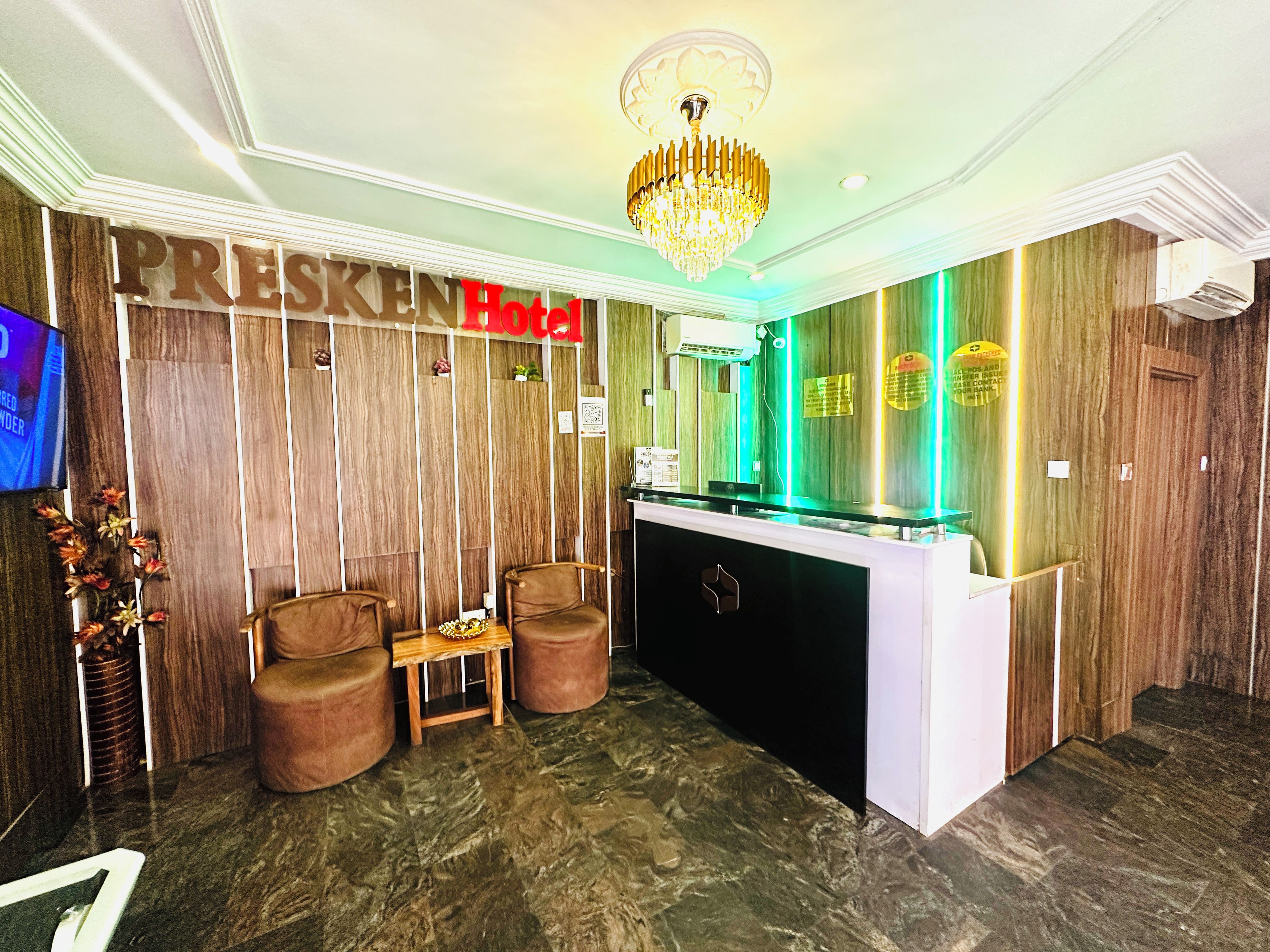 A hotel reception area with a wooden reception desk, two leather chairs, a chandelier, and a television on the left.