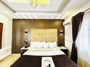 A hotel room with a large bed, a bedside telephone, a wall-mounted air conditioner, and a window with curtains. - Presken Freedom Way (Lekki)
