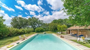 A rectangular swimming pool with clear water, surrounded by a tiled deck. There are lounge chairs and a small covered seating area with a roof.