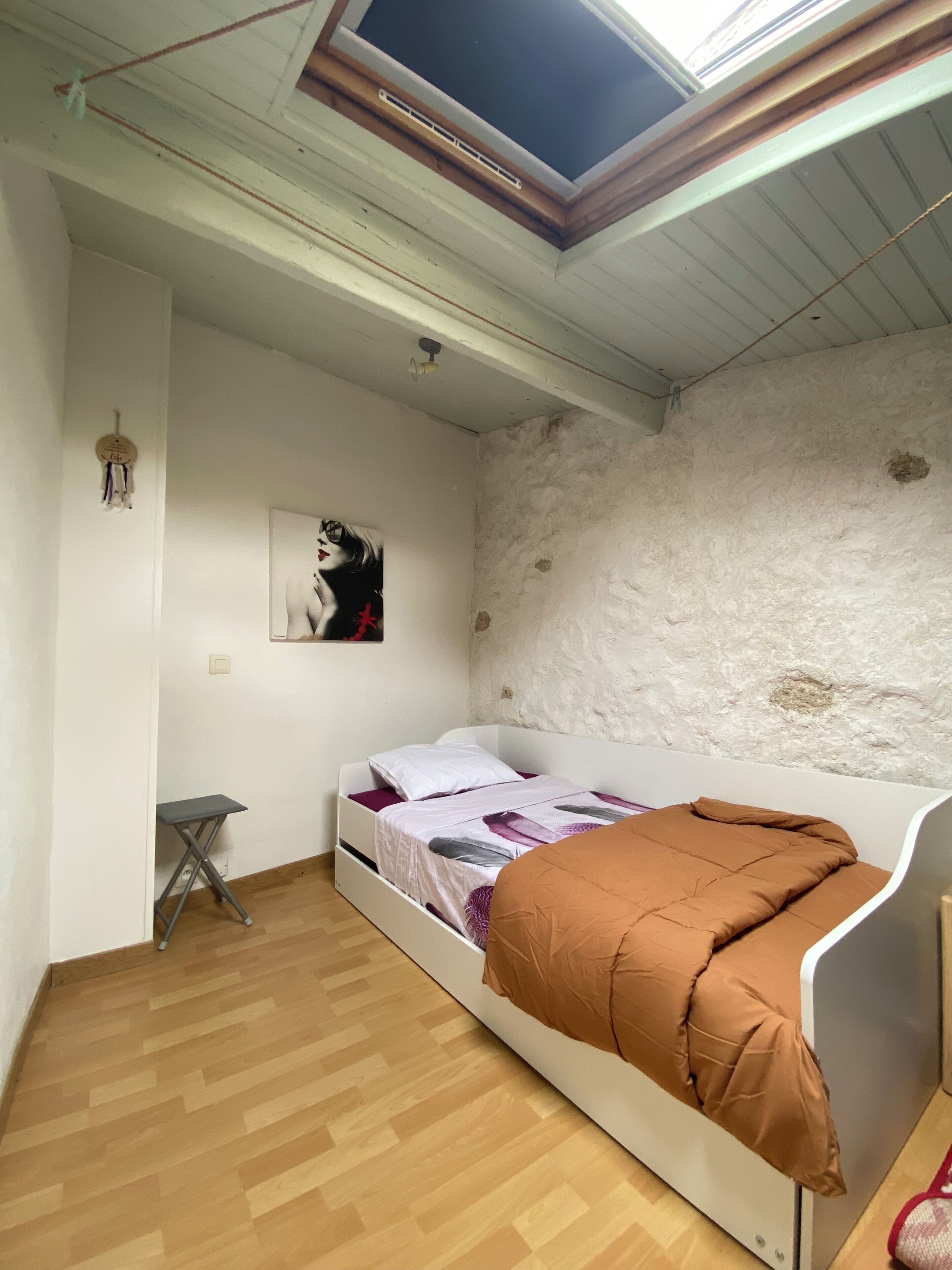 Una camera con un letto singolo, soffitto spiovente, pavimento in legno, parete bianca con una parte strutturata, un tavolino e una cornice con un'immagine.