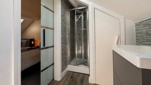 A compact room with a glass shower enclosure, a bed with patterned bedding, and a kitchenette area.