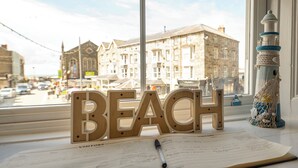 A window with a view of buildings and a wooden sign spelling 'BEACH' on a table with a notebook and pen.