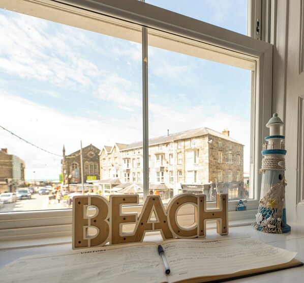 A window with a view of buildings and a wooden sign spelling 'BEACH' on a table with a notebook and pen. - Stunning Luxury Sea View 4 Bedroom Apartment - Glan Y Werydd House (Barmouth)
