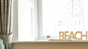 A room with a window, a sofa, a table with wine glasses and a bowl of fruit, and a sign reading 'BEACH' on the window sill.
