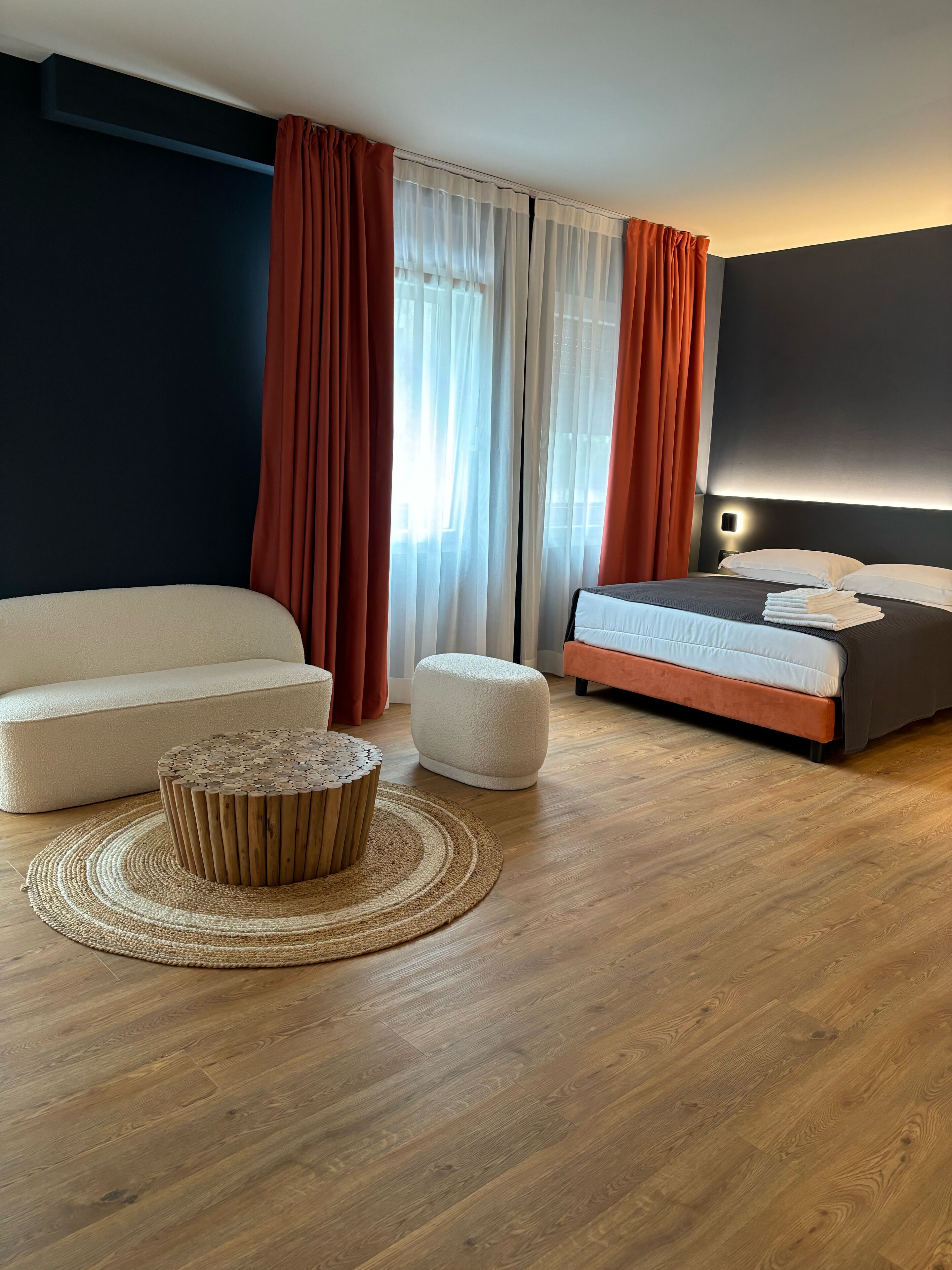 A modern hotel room with a large bed, a sofa, a round wooden coffee table, and a large window with curtains.
