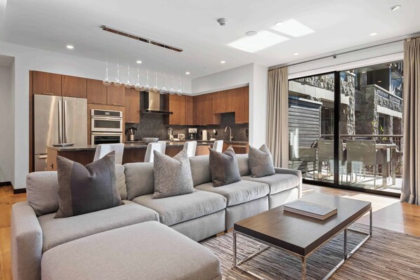 A modern living room with a sectional sofa, a coffee table, and a kitchen area with wooden cabinets and stainless steel appliances. - The Lion Vail - E209 (Vail)