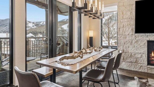 A dining area with a long table, chairs, a fireplace, and a view of the mountains. - The Lion Residences E503 (Vail)