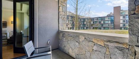 A balcony with a stone wall, a glass door leading to a room, and a view of a multi-story building.