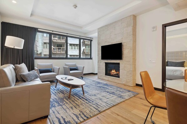 A modern living room with a fireplace, a flat-screen TV mounted on a stone wall, and a large window offering a view of the building's exterior. - The Lion Residences W301 (Vail)