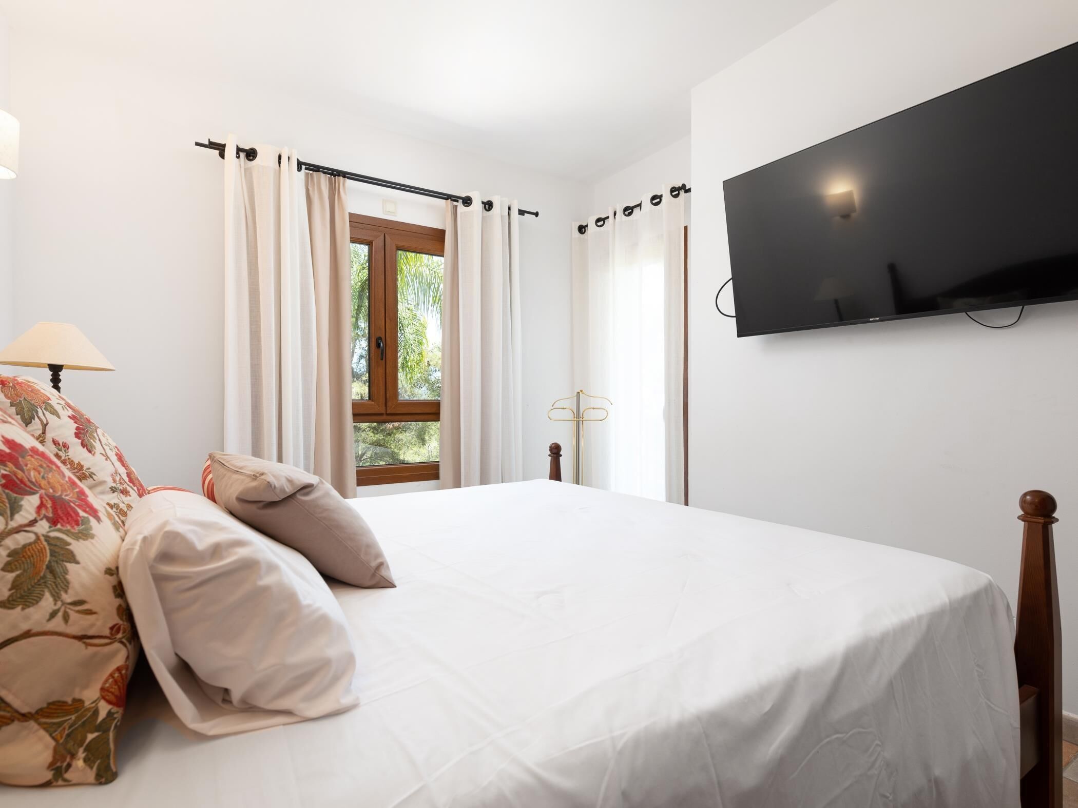 A bedroom with a bed, a TV mounted on the wall, a window with curtains, and a lamp.