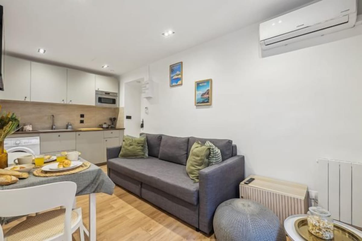 A modern living space with a grey sofa, a dining table with a vase of flowers, and a kitchen area with white cabinets.