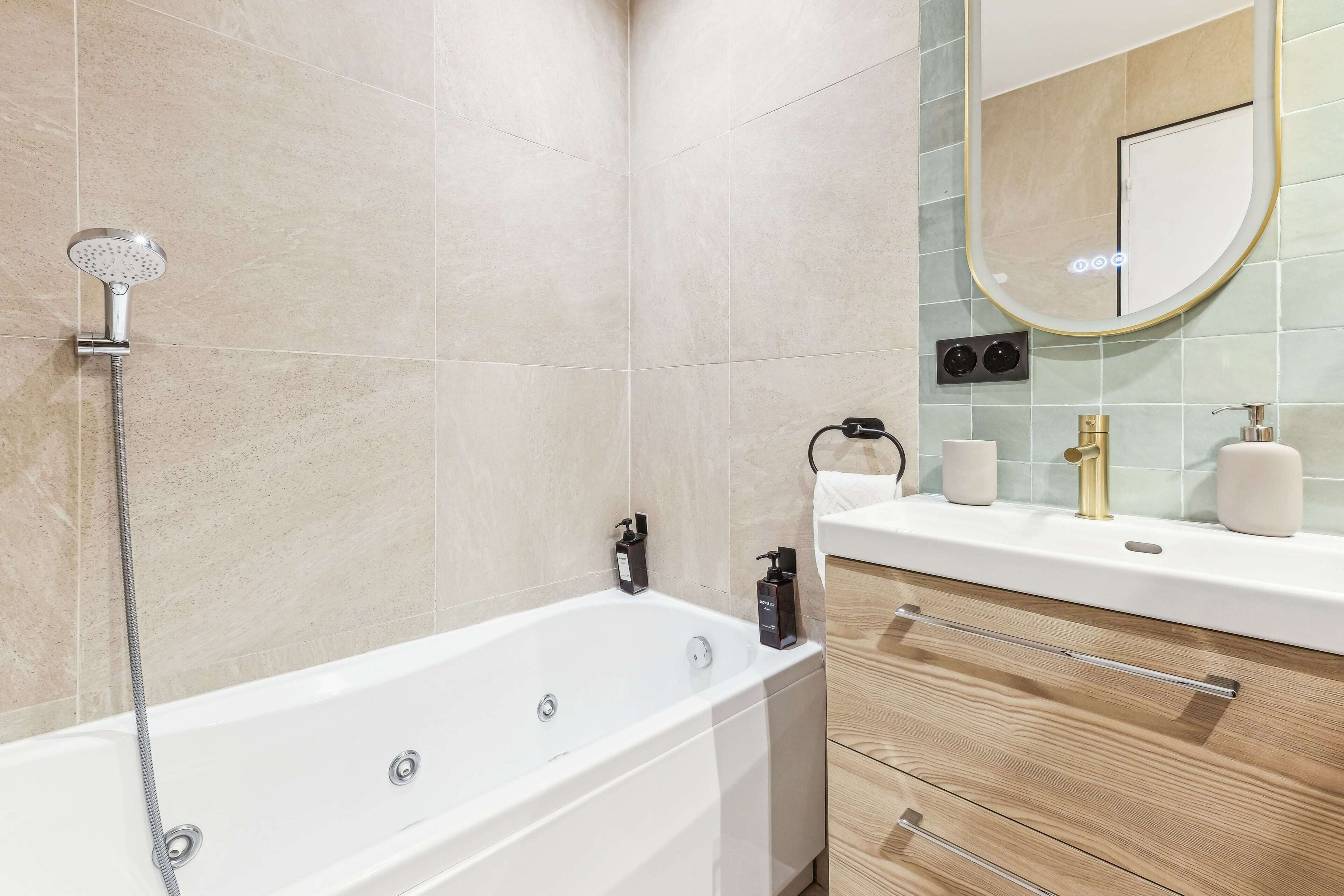 A modern bathroom with a white bathtub, a handheld showerhead, a wooden vanity with a gold faucet, and a mirror with a gold frame.