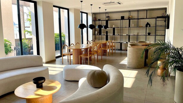Una sala moderna con sofá, reposapiés y mesa de centro, un comedor con mesa y sillas de madera, y una cocina con estantería negra.
