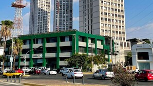 Un edificio moderno con fachada verde y varios pisos, situado en una zona urbana con otros edificios altos y un cielo azul despejado.