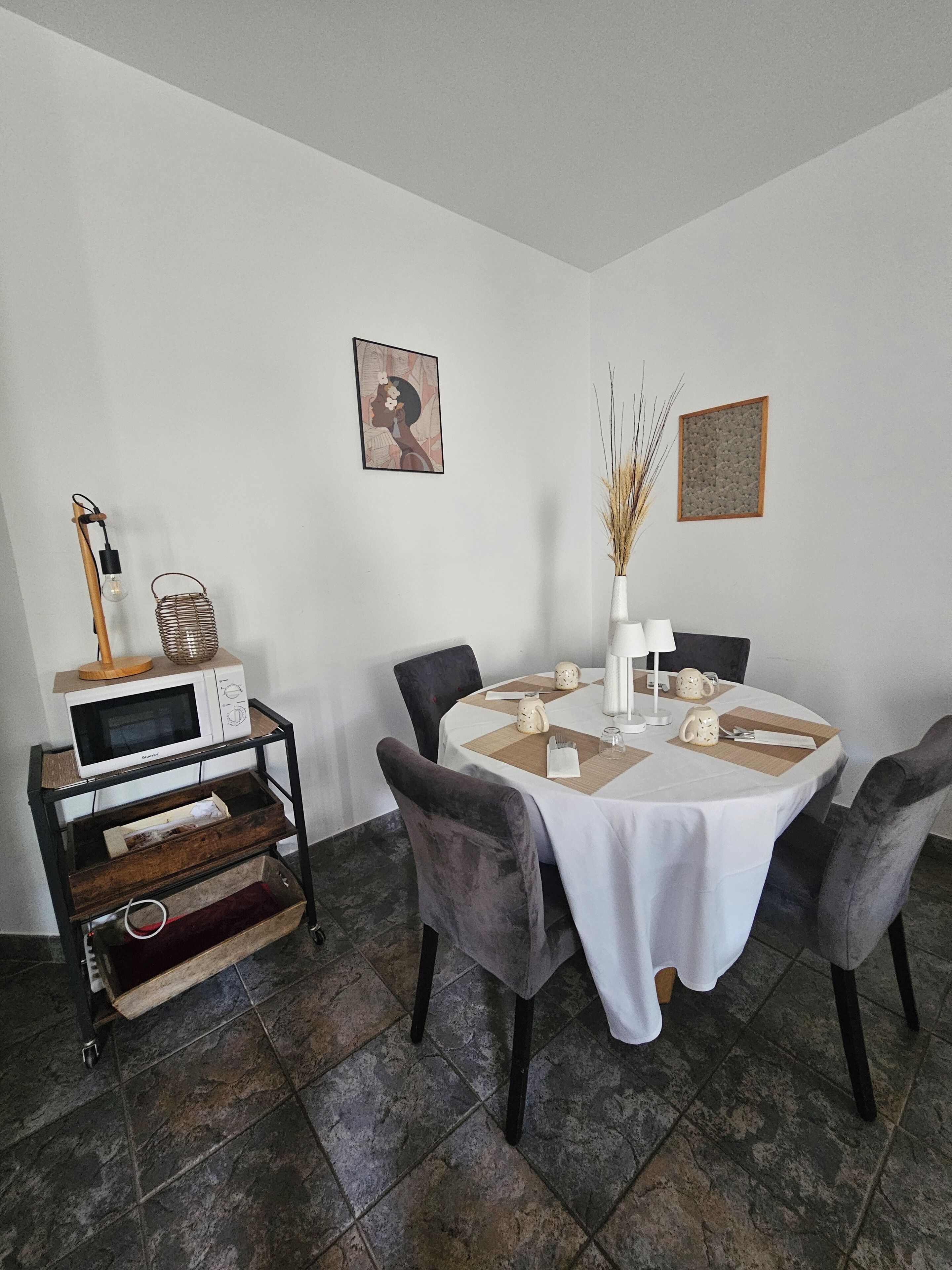 A dining area with a round table, four chairs, a white tablecloth, a microwave on a wooden cart, and two framed pictures on the wall.