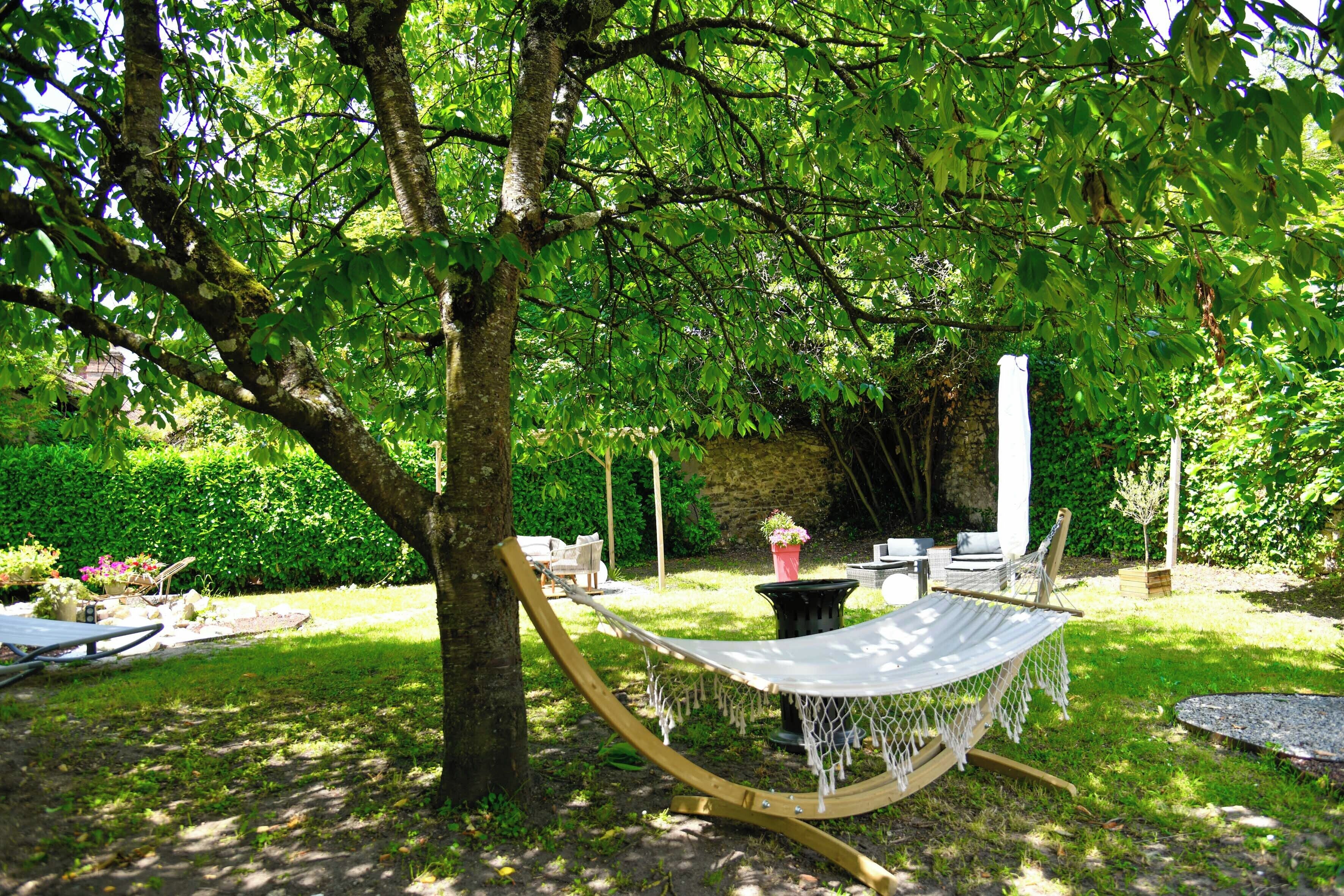 Una hamaca bajo un árbol en un jardín exuberante con un muro de piedra, muebles de exterior y una pequeña mesa.