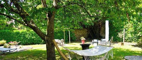 Una hamaca bajo un árbol en un jardín exuberante con un muro de piedra, muebles de exterior y una pequeña mesa.